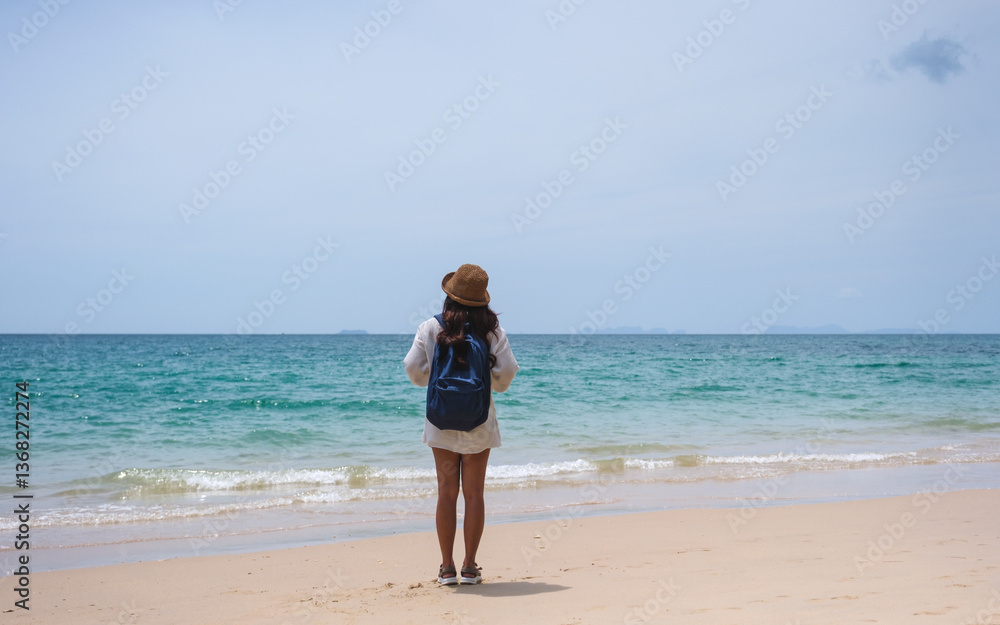 Rear view of a female traveler with hat and backpack standing on the beach by the sea Rear view of a female traveler with hat and backpack standing on the beach by the sea