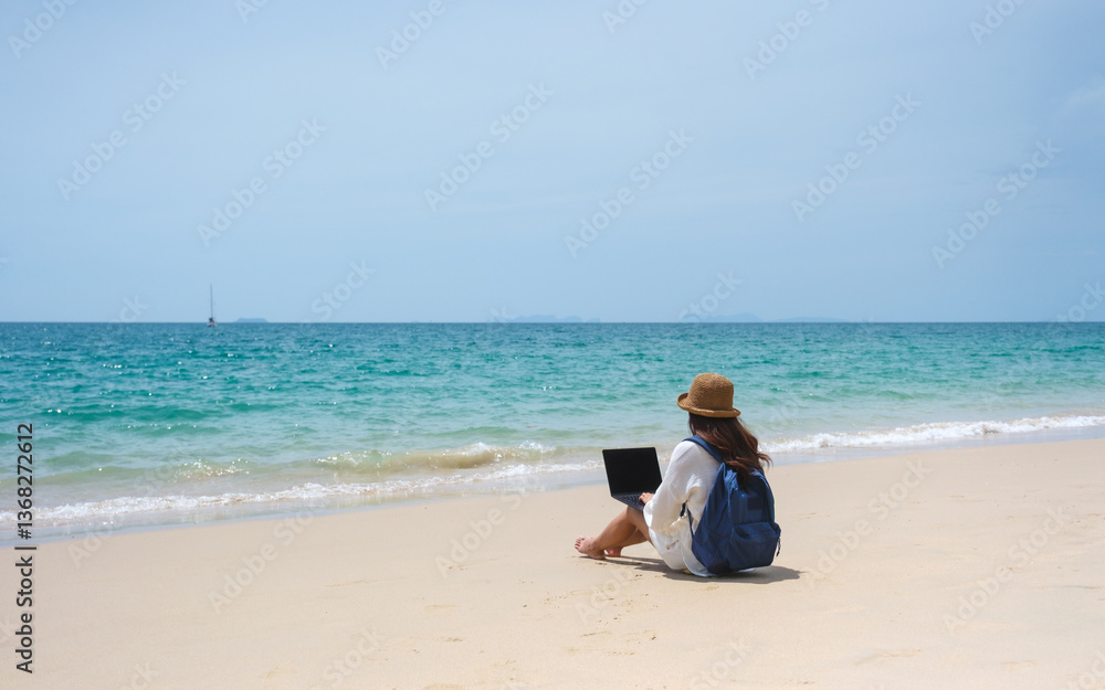 A female traveler using and working on laptop computer while sitting on the beach A female traveler using and working on laptop computer while sitting on the beach
