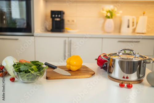 A fresh yellow bell pepper sits on a wooden cutting board in the kitchen. A knife is placed next to it, with other vegetables and a saucepan in the background.