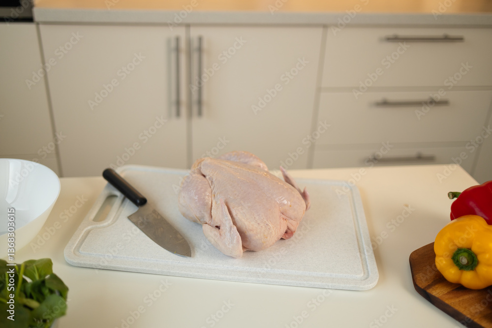 A raw whole chicken is placed on a white cutting board with a sharp kitchen knife next to it. Colorful bell peppers are also placed nearby on the kitchen counter.