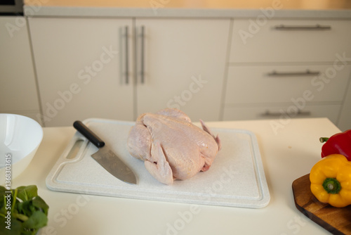 Obraz na płótnie A raw whole chicken is placed on a white cutting board with a sharp kitchen knife next to it. Colorful bell peppers are also placed nearby on the kitchen counter. w salonie