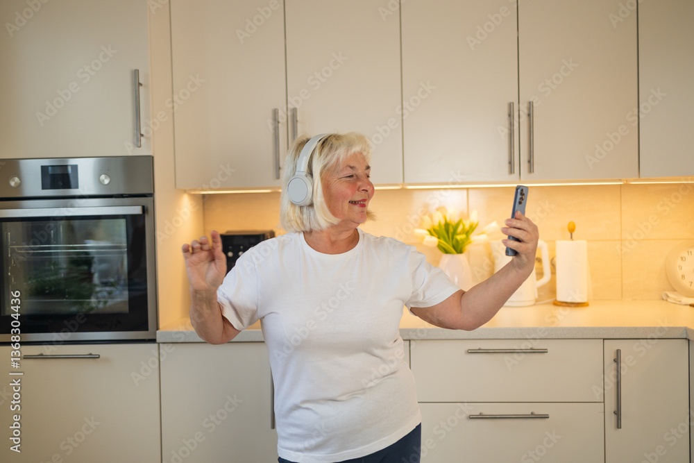 A senior woman stands in her kitchen, smiling and enjoying her favorite music. She listens to tunes on her phone, embracing the joy of the moment in her comfortable home setting