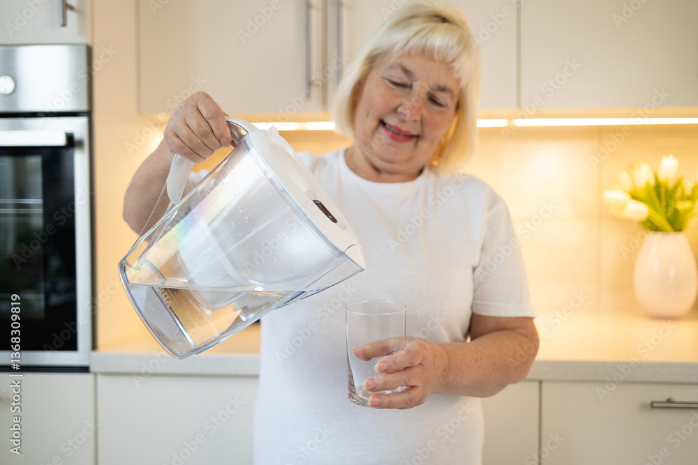 A happy senior woman holding a water pitcher and a glass in the kitchen, ready to pour herself a refreshing drink. This image reflects healthy habits and enjoying moments at home