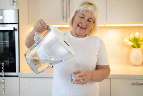 Obraz na płótnie A happy senior woman holding a water pitcher and a glass in the kitchen, ready to pour herself a refreshing drink. This image reflects healthy habits and enjoying moments at home w salonie