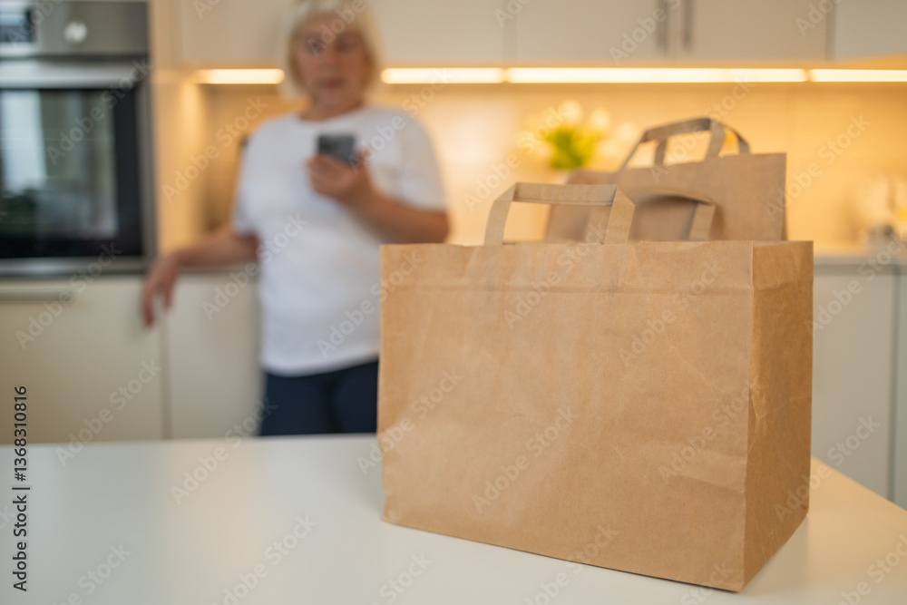 A senior woman stands in the kitchen, looking at her phone. In front of her are two paper grocery bags on the table, likely just delivered or picked up, ready for unpacking.