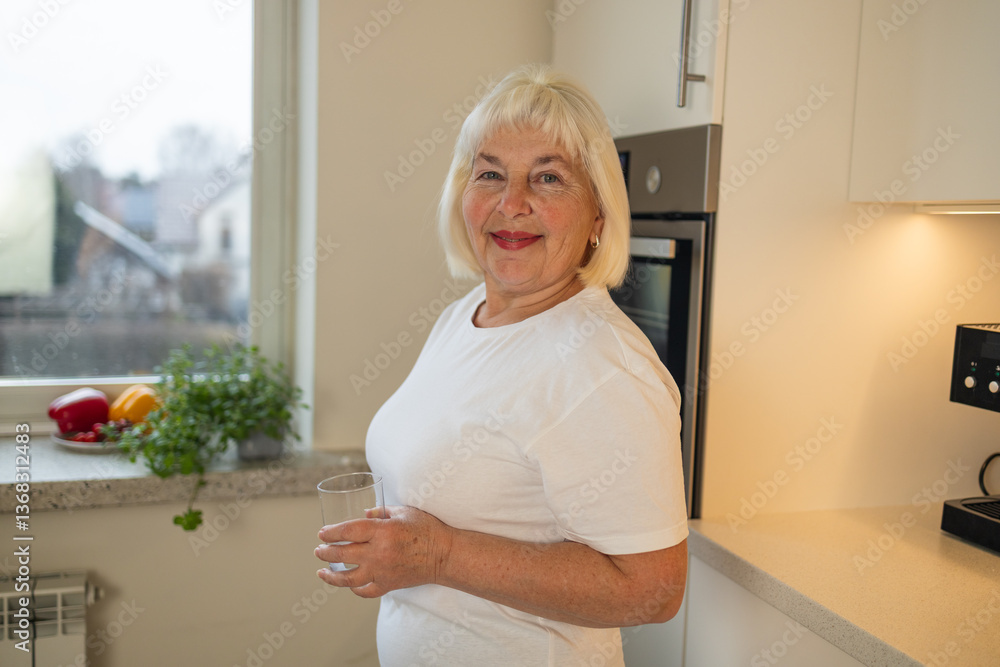 A joyful senior woman holding a glass of water in the kitchen, standing with a smile. This image captures the essence of healthy living and the pleasure of staying hydrated at home