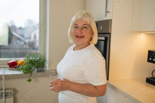 Obraz na płótnie A joyful senior woman holding a glass of water in the kitchen, standing with a smile. This image captures the essence of healthy living and the pleasure of staying hydrated at home w salonie