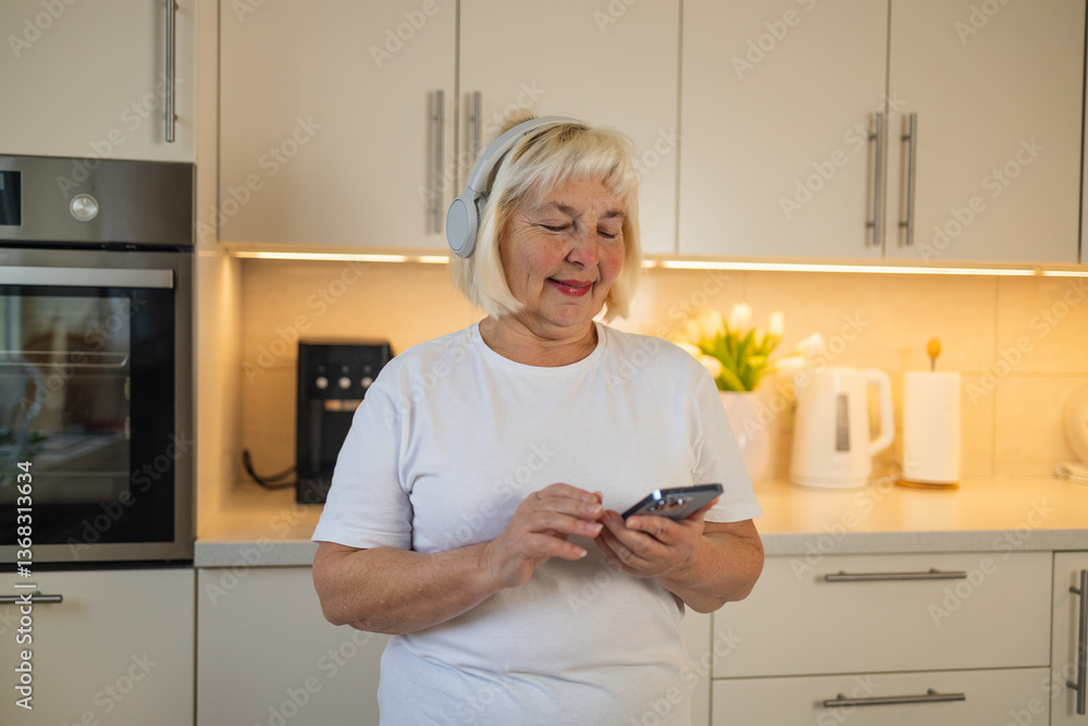 A senior woman stands in her kitchen, smiling and enjoying her favorite music. She listens to tunes on her phone, embracing the joy of the moment in her comfortable home setting