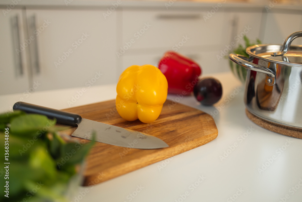 A fresh yellow bell pepper sits on a wooden cutting board in the kitchen. A knife is placed next to it, with other vegetables and a saucepan in the background.