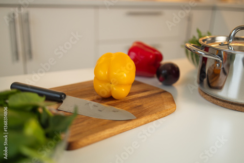 Obraz na płótnie A fresh yellow bell pepper sits on a wooden cutting board in the kitchen. A knife is placed next to it, with other vegetables and a saucepan in the background. w salonie