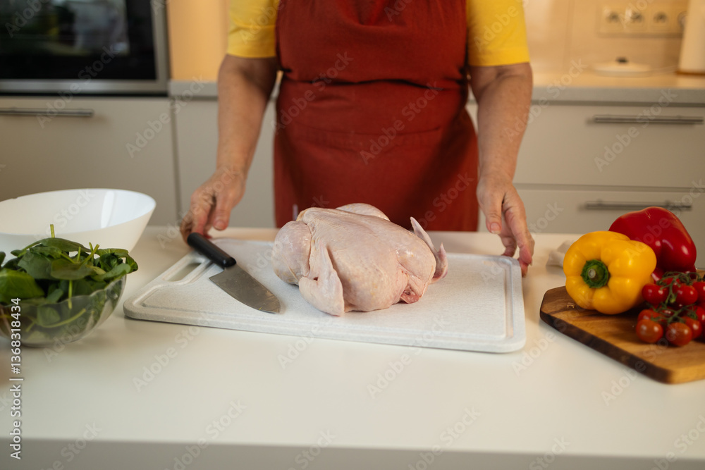 A woman in a yellow shirt and red apron is cutting raw chicken on a white cutting board in the kitchen. Colorful bell peppers and tomatoes are placed on the counter nearby.