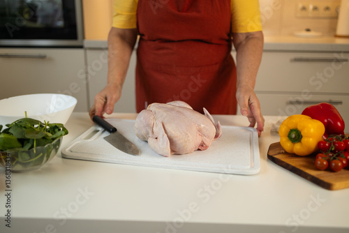 Obraz na płótnie A woman in a yellow shirt and red apron is cutting raw chicken on a white cutting board in the kitchen. Colorful bell peppers and tomatoes are placed on the counter nearby. w salonie