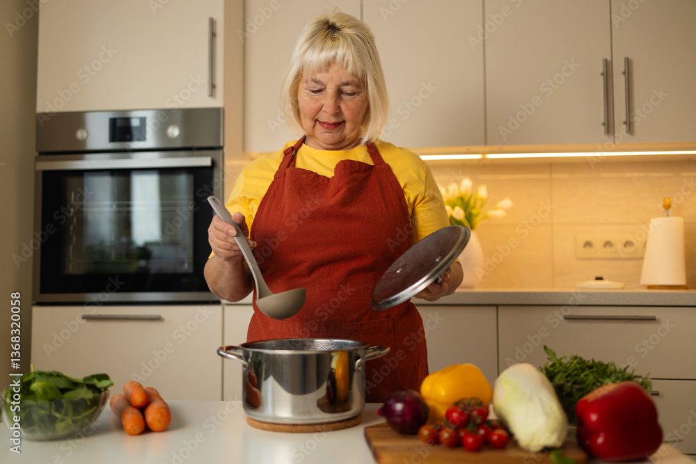A senior woman in a yellow apron is stirring vegetables in a pot on the kitchen counter, with fresh carrots, tomatoes, and other ingredients around her.