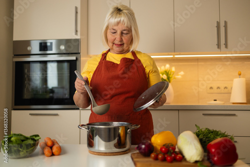 Obraz na płótnie A senior woman in a yellow apron is stirring vegetables in a pot on the kitchen counter, with fresh carrots, tomatoes, and other ingredients around her. w salonie