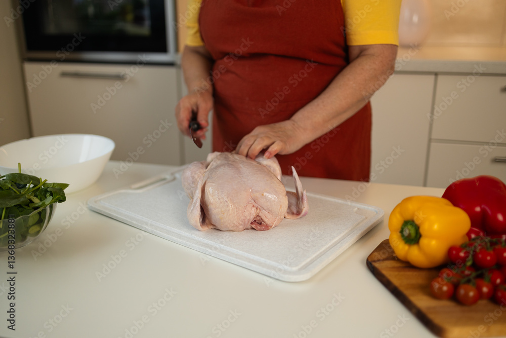 A woman in a yellow shirt and red apron is cutting raw chicken on a white cutting board in the kitchen. Colorful bell peppers and tomatoes are placed on the counter nearby.