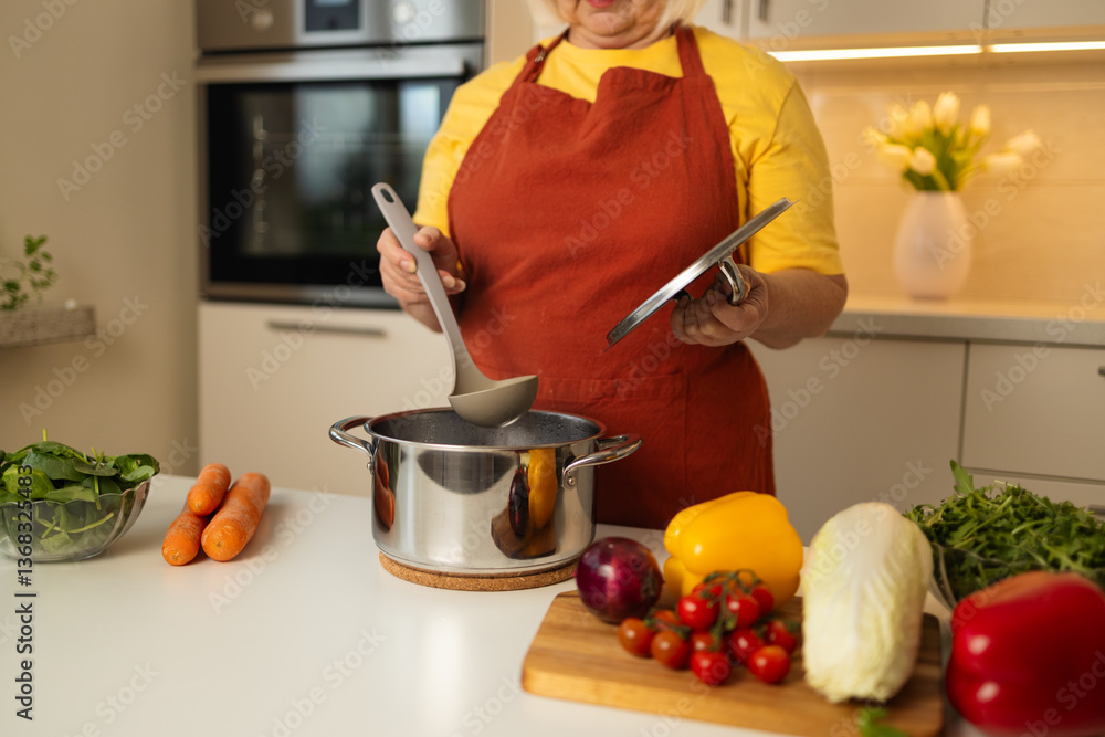 A senior woman in a yellow apron is stirring vegetables in a pot on the kitchen counter, with fresh carrots, tomatoes, and other ingredients around her.