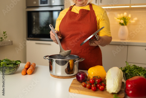 A senior woman in a yellow apron is stirring vegetables in a pot on the kitchen counter, with fresh carrots, tomatoes, and other ingredients around her.