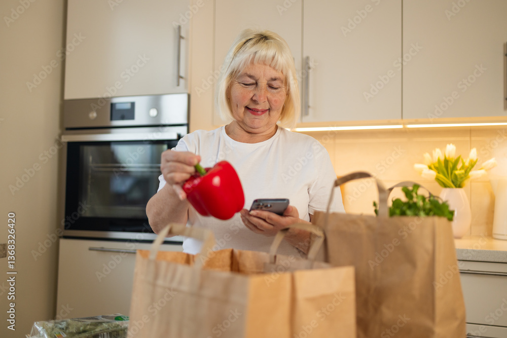 An elderly woman is holding a red bell pepper and looking at her phone while unpacking groceries in her modern kitchen. She smiles as she checks something on her phone.