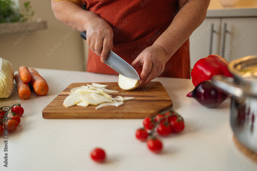 A person slices onion on a wooden cutting board in a bright kitchen. Various fresh vegetables, including tomatoes, carrots, and bell peppers, are placed around for meal preparation