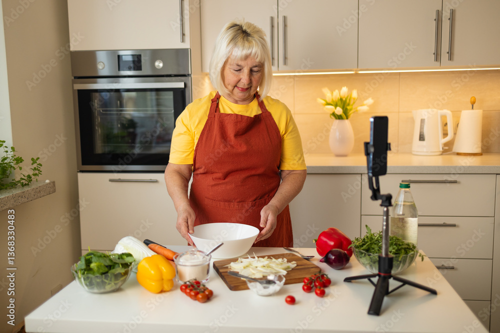 An elderly woman in a yellow shirt and apron prepares food in a bright kitchen. Fresh vegetables like tomatoes, peppers, and spinach are on the table. A camera is set up for filming her cooking
