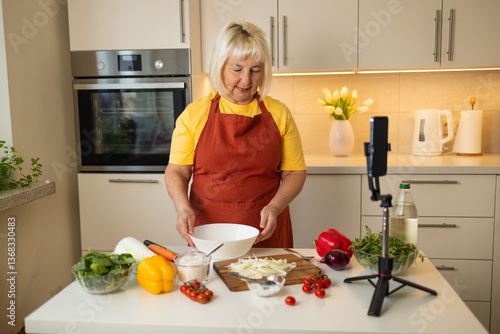 Obraz na płótnie An elderly woman in a yellow shirt and apron prepares food in a bright kitchen. Fresh vegetables like tomatoes, peppers, and spinach are on the table. A camera is set up for filming her cooking w salonie