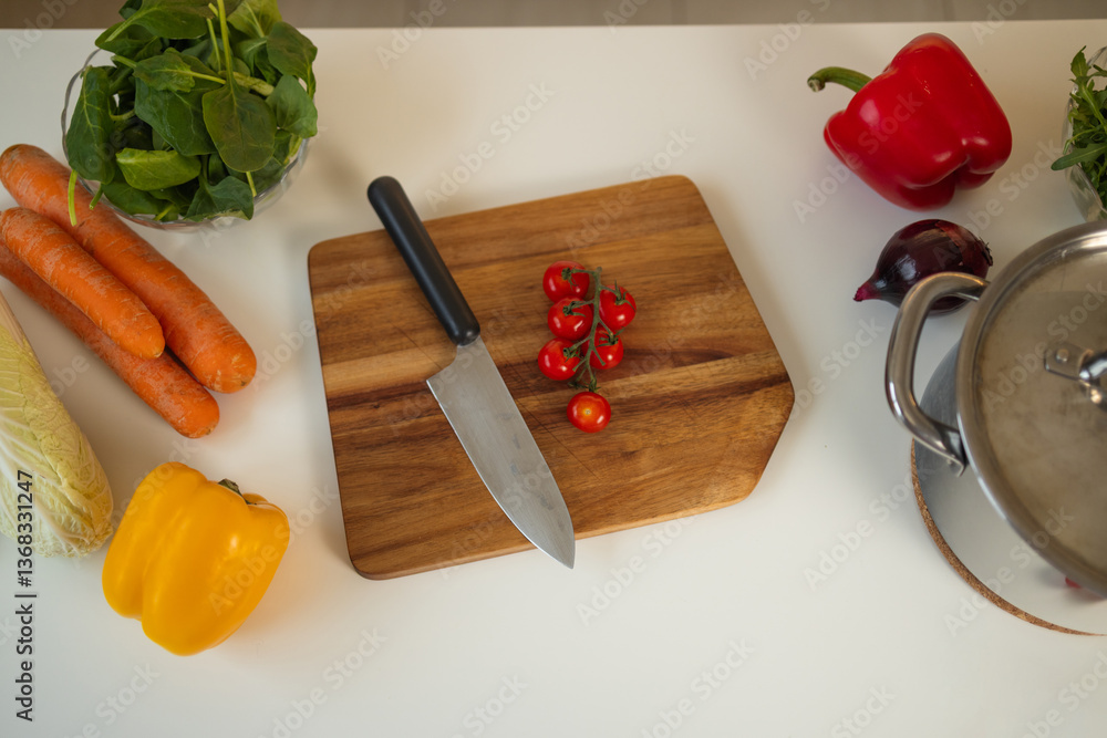 Fresh cherry tomatoes are sliced on a wooden cutting board in a bright kitchen. A sharp knife is used, and colorful vegetables like carrots, bell pepper, and spinach are nearby.
