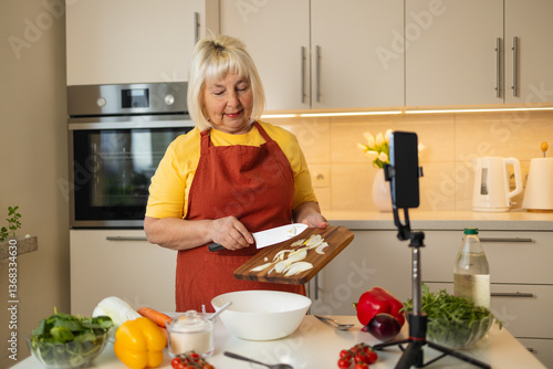 Obraz na płótnie A senior woman in a yellow shirt and brown apron mixes ingredients in a bowl while preparing a healthy meal in a bright kitchen. Fresh vegetables like peppers, tomatoes, and spinach are on the table. w salonie