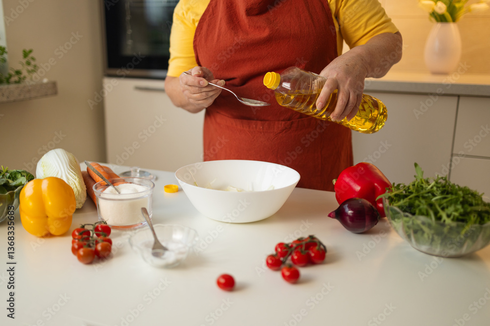 A senior woman in a yellow shirt and brown apron pours cooking oil into a bowl while preparing a dish in a modern kitchen. Fresh vegetables are laid out on the counter for meal preparation