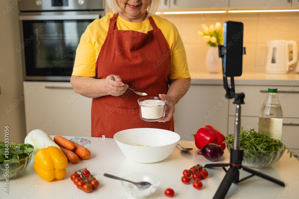 A senior woman in a yellow shirt and brown apron adds sugar to a bowl while preparing a meal in a well-organized kitchen. Fresh vegetables and kitchen tools are visible on the countertop