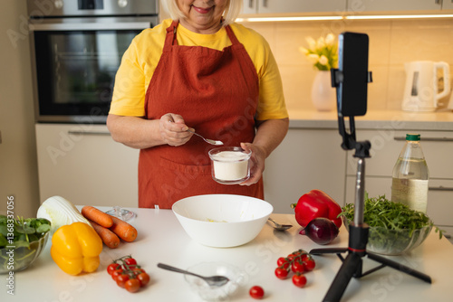 Obraz na płótnie A senior woman in a yellow shirt and brown apron adds sugar to a bowl while preparing a meal in a well-organized kitchen. Fresh vegetables and kitchen tools are visible on the countertop w salonie