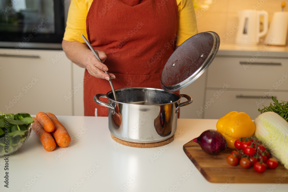 A senior woman in a yellow apron is stirring vegetables in a pot on the kitchen counter, with fresh carrots, tomatoes, and other ingredients around her.