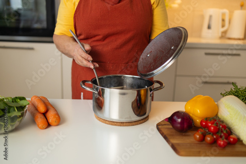 Obraz na płótnie A senior woman in a yellow apron is stirring vegetables in a pot on the kitchen counter, with fresh carrots, tomatoes, and other ingredients around her. w salonie