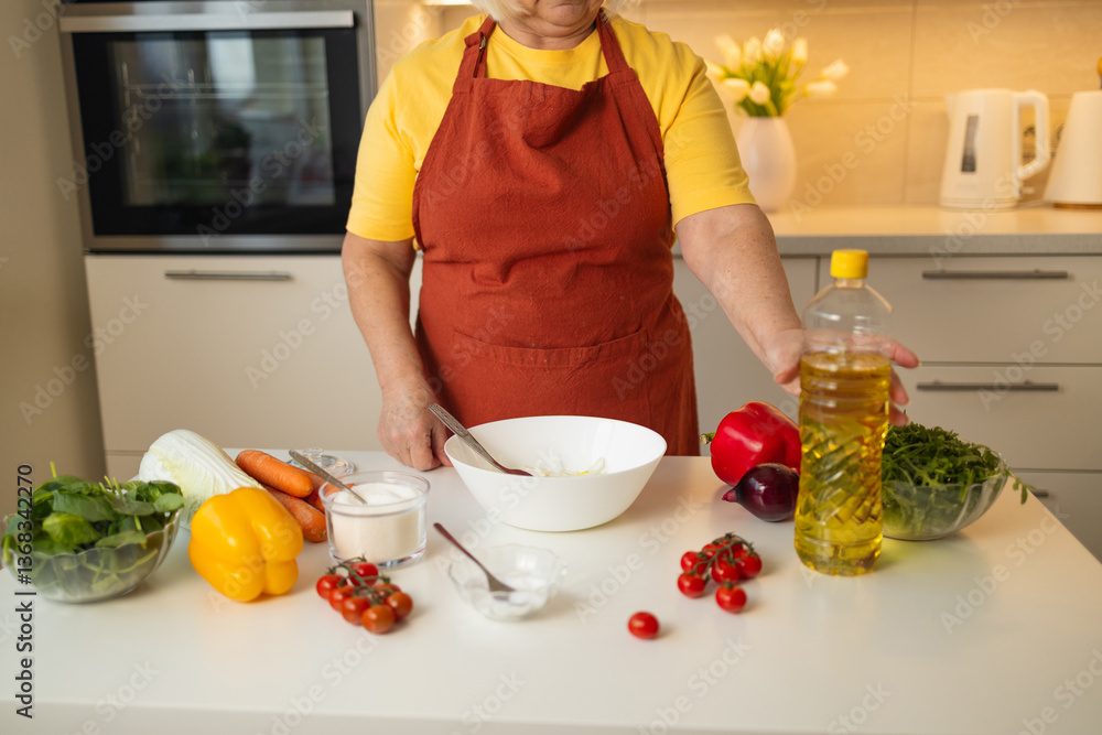 A senior woman in a yellow shirt and brown apron pours cooking oil into a bowl while preparing a dish in a modern kitchen. Fresh vegetables are laid out on the counter for meal preparation