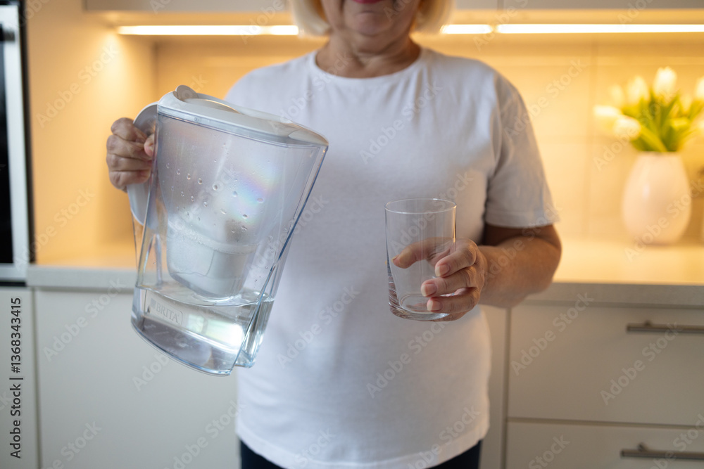 A happy senior woman holding a water pitcher and a glass in the kitchen, ready to pour herself a refreshing drink. This image reflects healthy habits and enjoying moments at home