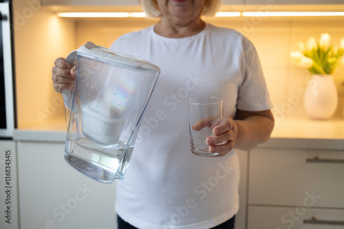 A happy senior woman holding a water pitcher and a glass in the kitchen, ready to pour herself a refreshing drink. This image reflects healthy habits and enjoying moments at home