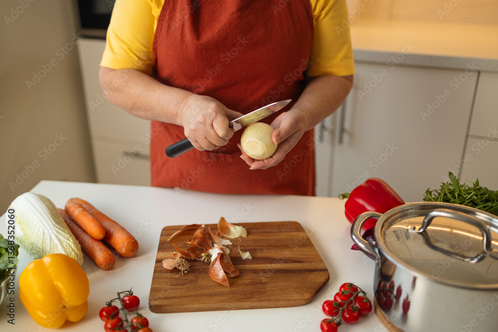 A woman in a yellow shirt and apron is peeling an onion in the kitchen. She is preparing ingredients for a meal while surrounded by fresh vegetables like carrots, tomatoes, and bell peppers