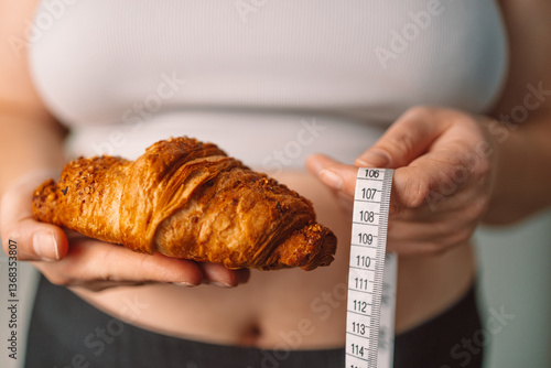 Obraz na płótnie A woman holding a croissant in one hand and a measuring tape in the other, reflecting the challenge of balancing indulgent food with health and wellness goals. Concept of dieting and self-control w salonie