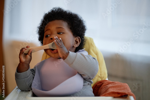 Medium shot of hungry Black baby girl wearing silicone bib biting spoon during mealtime while sitting in high chair in kitchen with warm tone, copy space