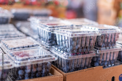 Multiple clear square plastic containers of organic blueberries for sale at a farmer's market. The packages are stacked in cardboard trays on display. The fresh cultivated berries are bright blue.