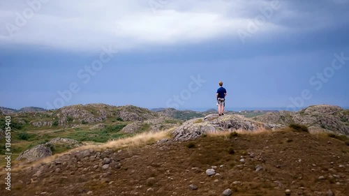 A young boy stands on a rocky outcrop, gazing at a dramatic sky over rolling hills