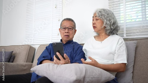 Elderly couple sitting on sofa, smiling and looking at smartphone in cozy living room, sharing emotional and curious moments.