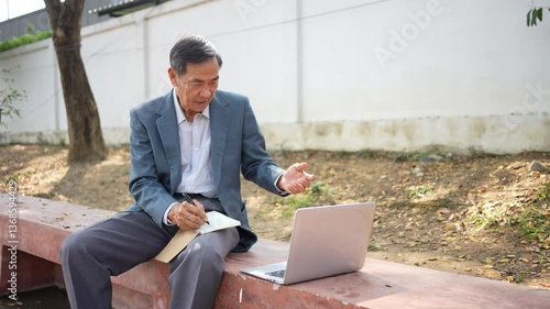 Elderly man in suit sitting outdoors on bench, using laptop and holding notebook, senior working focused in park setting.