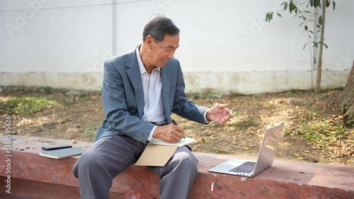 Elderly man in suit sits outdoors on bench, writing in notebook while using laptop, appearing focused and thoughtful.