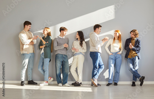 Group of young people casually chatting while leaning against gray wall in modern space. Close friends or colleagues engaged in friendly conversation, enjoying social interaction and connection.