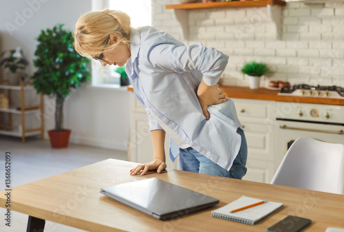 Tired blonde woman standing in kitchen, holding lower back in pain, leaning on wooden table. Unhappy stressed young lady suffering from spasm or pinched nerve after long work or sedentary lifestyle.