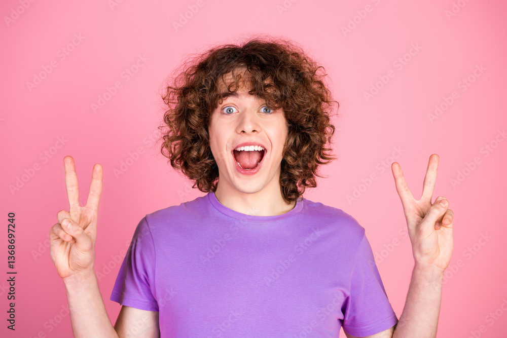 Joyful young man with curly hair posing with peace signs on a pink background, expressing excitement and positivity Joyful young man with curly hair posing with peace signs on a pink background, expressing excitement and positivity