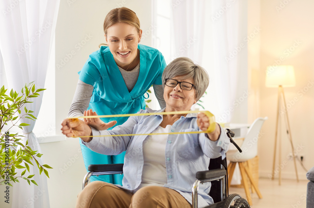 Fototapeta premium Supportive caregiver helping to disabled old woman with doing physical exercises and rehabilitation, stretching resistance band with hands, cannot walk, posing in wheelchair. Caregiving concept