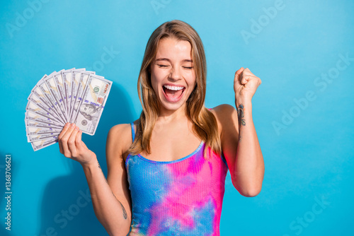 Canvas-taulu Excited woman celebrating success holding dollar bills against a vibrant blue ba