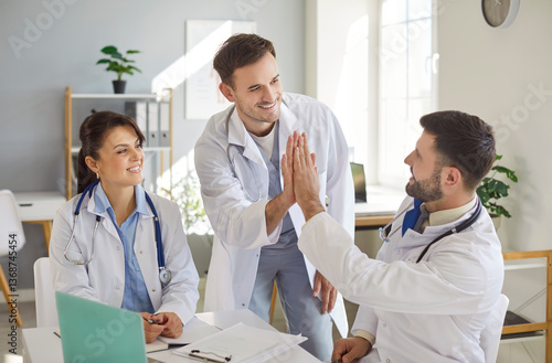 Three cheerful doctors in white coats gathered around table in bright medical office. Smiling physician giving high five to his colleague, team celebrating successful collaboration and achievement.