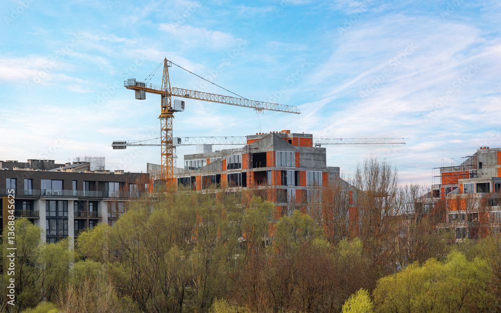 A modern residential complex under construction, featuring concrete and brick structures with large glass windows, surrounded by trees, with cranes towering above against a clear blue sky.